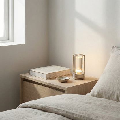 Nighstand with a book, small bowl, and lit candle next to a bed in a softly lit room.