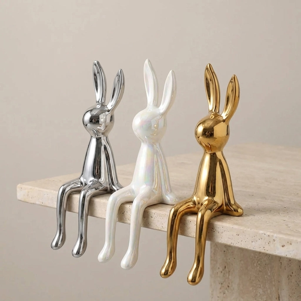 Three decorative rabbit figurines in gold, silver, and white on a wooden shelf.