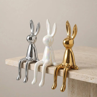Three decorative rabbit figurines in gold, silver, and white on a wooden shelf.