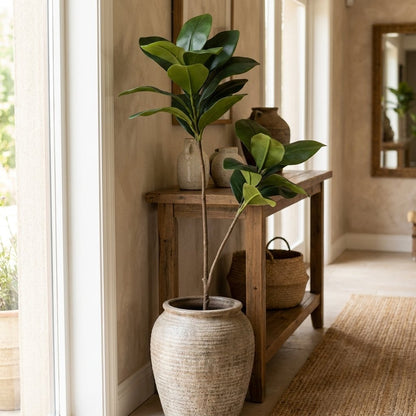 Two faux magnolia tree stems in a ribbed terracotta urn on a rustic console table beside a mirror and woven baskets