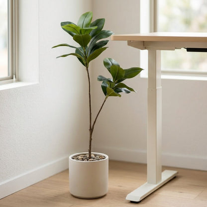 Two faux magnolia tree stems in a white cylindrical pot topped with decorative pebbles, beside a white standing desk