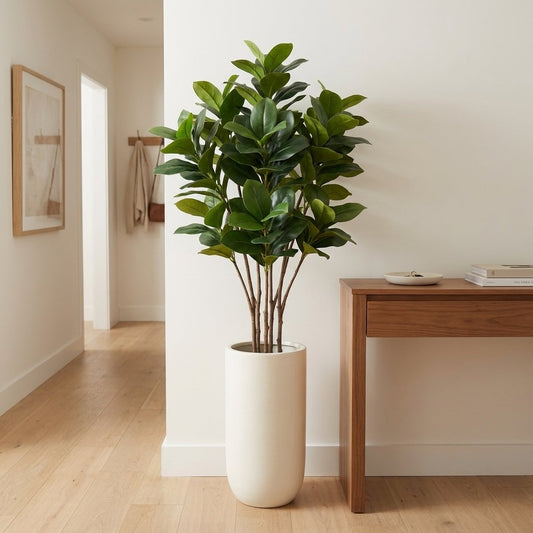 Bushy faux magnolia tree in a tall white tapered planter in a hallway corner, beside a wood console table with books and framed art