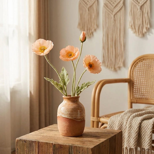Four faux poppy stems with three peach-apricot blooms and a bud in a cream and terracotta ceramic vase on a rustic wood table, with macrame wall hangings and a rattan chair behind