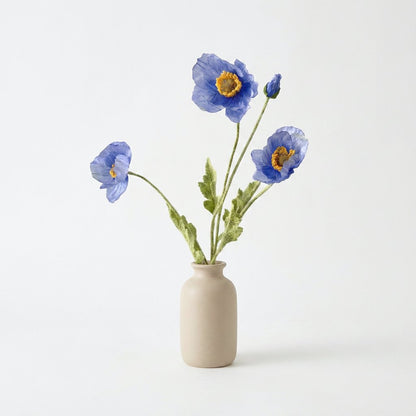 Four faux poppy stems with three periwinkle blue blooms with amber centers and a blue bud in a small cream ceramic vase on a plain light backdrop
