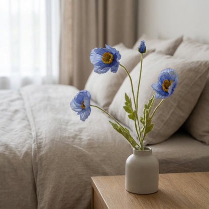Four faux poppy stems with three periwinkle blue blooms and a bud in a small cream ceramic bottle vase on a light oak nightstand beside a bed with beige linen bedding