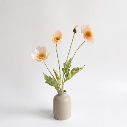 Four faux poppy stems with three very pale peach blush blooms and a bud in a small speckled taupe stoneware jar on a plain light backdrop