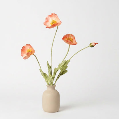 Four faux poppy stems with three salmon-coral blooms with cream-pink centers and a coral bud in a matte tan ceramic jar on a plain light backdrop