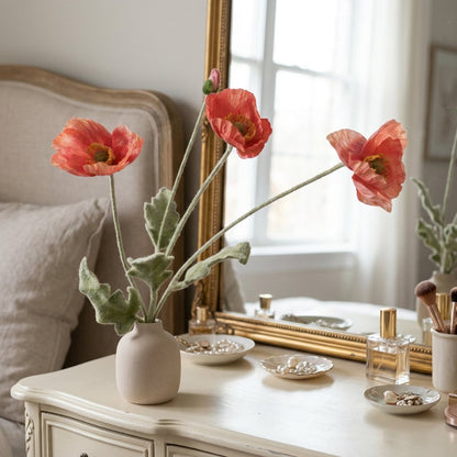 Four faux poppy stems with three red-orange coral blooms and a bud in a speckled cream vase on a cream dresser with a gold-framed mirror, a perfume bottle and jewelry dishes