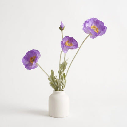 Four faux poppy stems with three medium lavender-violet blooms and a purple bud in a cream ceramic jar vase on a plain light backdrop