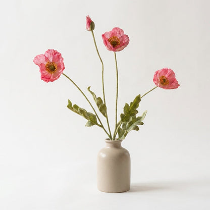 Four faux poppy stems with three rose-pink blooms with lighter edges and a pink bud in a greige ceramic bottle vase on a plain warm off-white backdrop