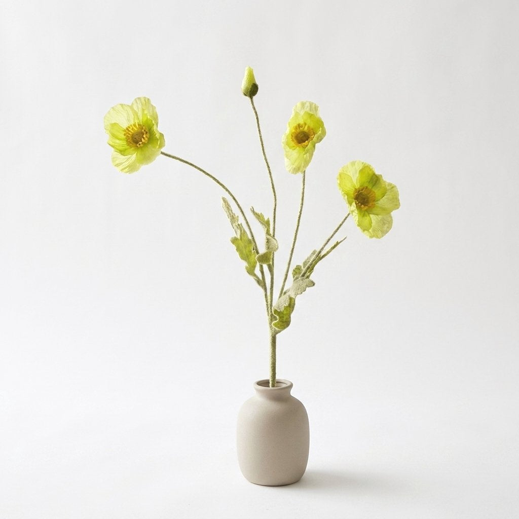 Four faux poppy stems with three pale lime yellow-green blooms and a lime bud in a matte taupe ceramic jar on a plain light backdrop