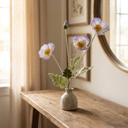 Four faux poppy stems with three white blooms edged in lavender and a lavender bud in a speckled cream vase on a light wood console table beneath a round mirror