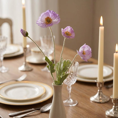 Four faux poppy stems with three lilac blooms and a bud in a tall white ceramic bud vase on a dining table set with gold-rimmed plates, crystal glasses and lit taper candles