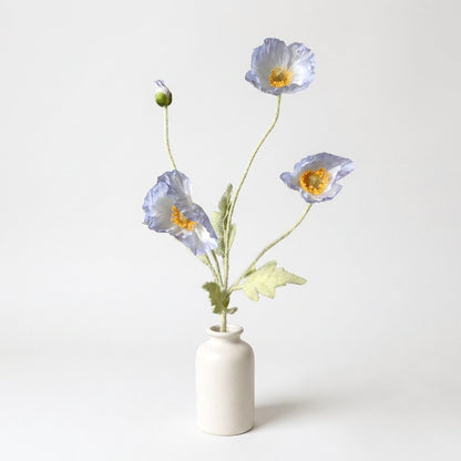 Four faux poppy stems with three pale periwinkle blooms with near-white centers and a bud in a matte white ceramic jar on a plain light backdrop