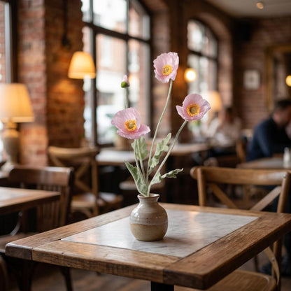 Four faux poppy stems with three light pink blooms and a bud in a small speckled ceramic vase on a cafe table with a white marble inset top and a blurred brick-walled cafe behind
