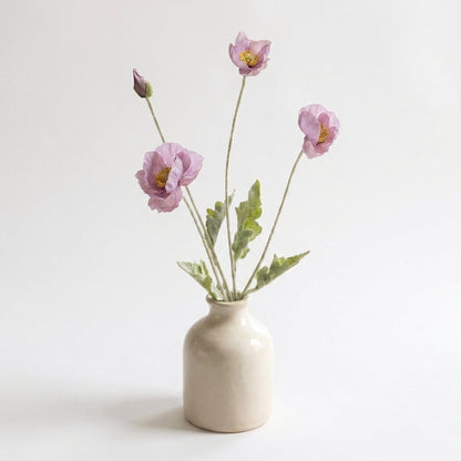 Four faux poppy stems with three dusty mauve blooms and a mauve-purple bud in a glossy cream ceramic jar vase on a plain light backdrop