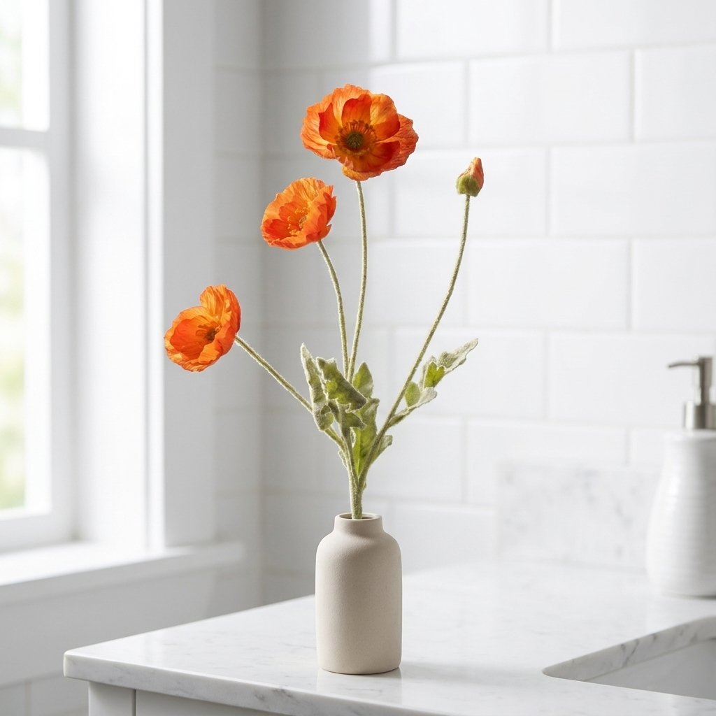 Four faux poppy stems with three orange blooms and a bud in a matte cream bottle vase on a white marble bathroom counter against white subway tile