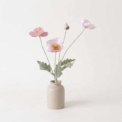 Four faux poppy stems with three soft pastel pink blooms and a bud in a beige ceramic jar vase on a plain off-white backdrop