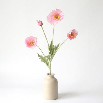 Four faux poppy stems with three medium rose-pink blooms and a deeper pink bud in a beige ceramic bottle vase on a plain light backdrop