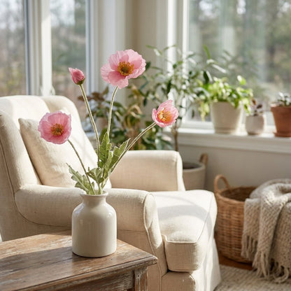Four faux poppy stems with three pink blooms and a bud in a glossy cream vase on a rustic wood table in a sunroom with a linen armchair and potted plants on the windowsill