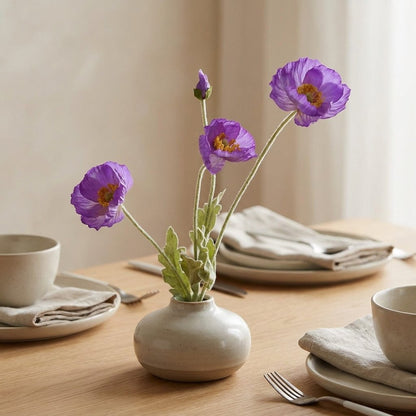 Four faux poppy stems with three violet-purple blooms and a bud in a speckled cream vase on an oak dining table among beige plates, linen napkins and silverware