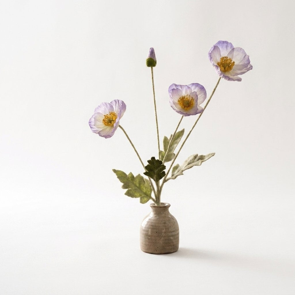 Four faux poppy stems with three white blooms fading to lavender at the edges and a lavender bud in a small speckled stoneware pot on a plain light backdrop