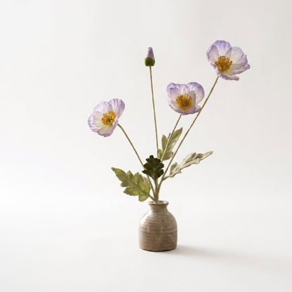Four faux poppy stems with three white blooms fading to lavender at the edges and a lavender bud in a small speckled stoneware pot on a plain light backdrop