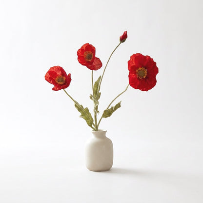 Four faux poppy stems with three saturated red blooms with dark centers and a red bud in a cream ceramic bottle vase on a plain light backdrop