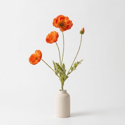 Four faux poppy stems with three orange blooms with deeper red-orange shading and a bud in a cream ceramic vase on a plain light gray backdrop