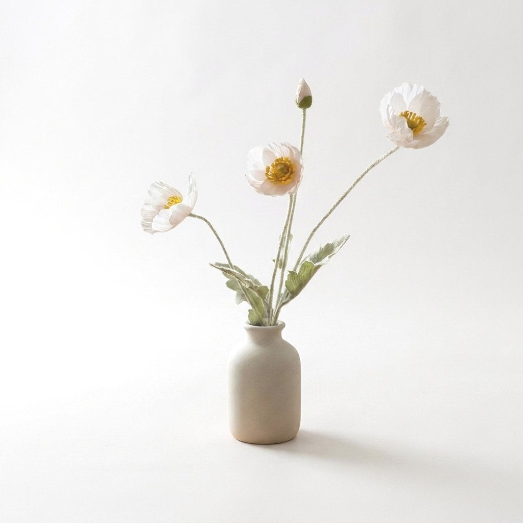 Four faux poppy stems with three pale blush blooms and a bud in a matte cream ceramic bottle vase on a plain light backdrop