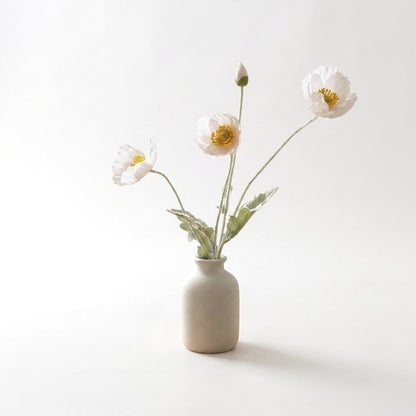 Four faux poppy stems with three pale blush blooms and a bud in a matte cream ceramic bottle vase on a plain light backdrop