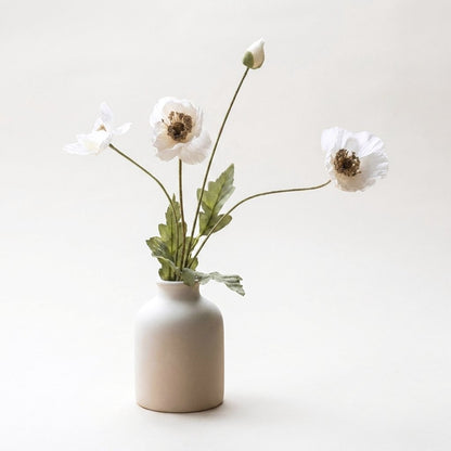 Four faux poppy stems with three creamy white ruffled blooms with dark brown centers and a white bud in a matte cream ceramic bottle vase on a plain warm off-white backdrop