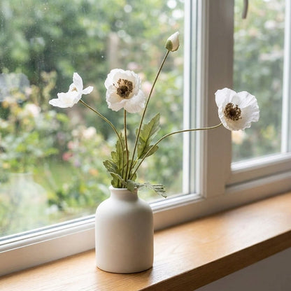 Four faux poppy stems with three white blooms with dark centers and a bud in a small cream ceramic vase on a wooden windowsill in front of a window onto a garden