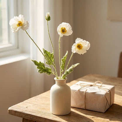 Four faux poppy stems with three white blooms and a bud in a cream ceramic bottle vase on a wood table beside a kraft-wrapped gift box tied with twine and a tag reading FOR YOU.