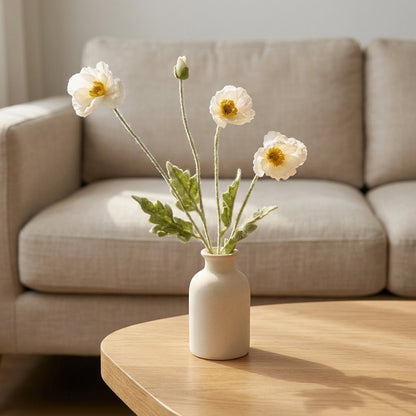 Four faux poppy stems with three white blooms and a bud in a small cream ceramic bottle vase on a light oak coffee table in front of an oatmeal sofa