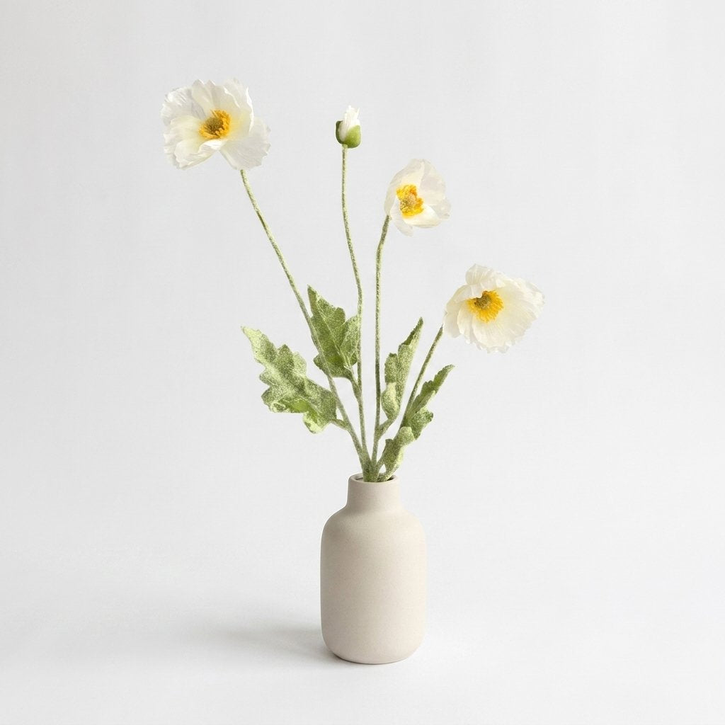 Four faux poppy stems with three white ruffled blooms with yellow-orange centers and a white bud in a matte cream ceramic bottle vase on a plain light backdrop