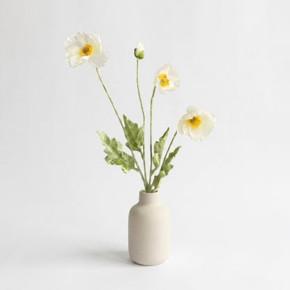Four faux poppy stems with three white ruffled blooms with yellow-orange centers and a white bud in a matte cream ceramic bottle vase on a plain light backdrop