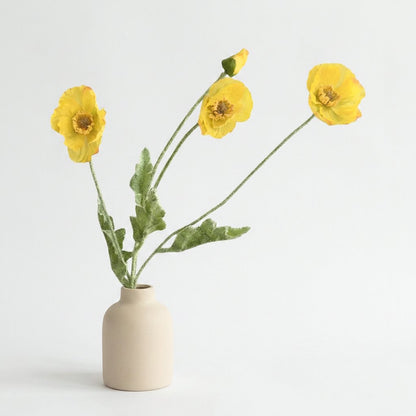 Four faux poppy stems with three bright yellow blooms and a bud in a matte cream ceramic bottle vase on a plain light backdrop