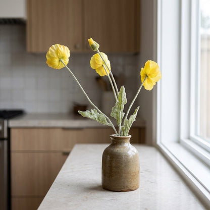 Four faux poppy stems with three yellow blooms and a bud in a tan and olive glazed jar on a cream stone kitchen counter beside a window, with light wood cabinets and a tile backsplash behind