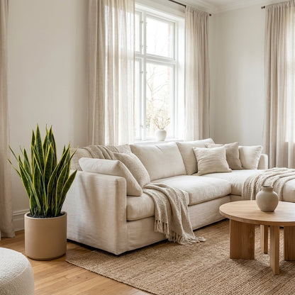 Faux snake plant in a tan planter beside a cream sectional sofa and round wood coffee table