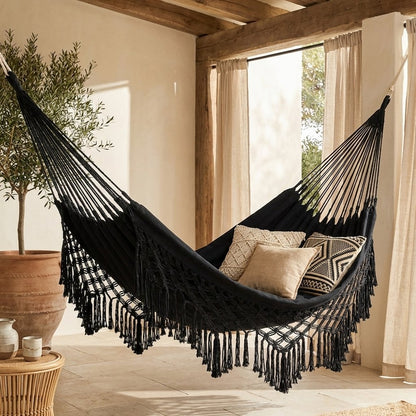 Black fringe macrame hammock with three cushions hung in a covered patio with wood beams and cream linen curtains, beside a potted olive tree and a round rattan side table
