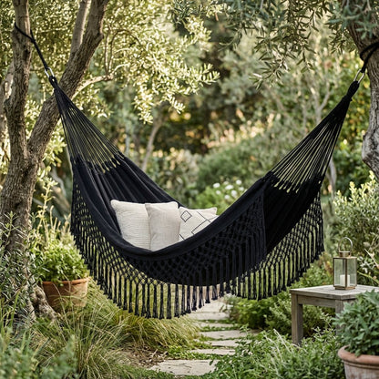 Black fringe macrame hammock with three cushions hung between two olive tree trunks in a garden, beside a wood side table holding a candle lantern