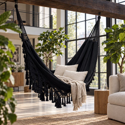 Black fringe macrame hammock hung from a rough wooden ceiling beam in a loft with a window wall, holding two cream pillows and a cream chunky-knit throw, with fiddle-leaf figs and a round wood side table