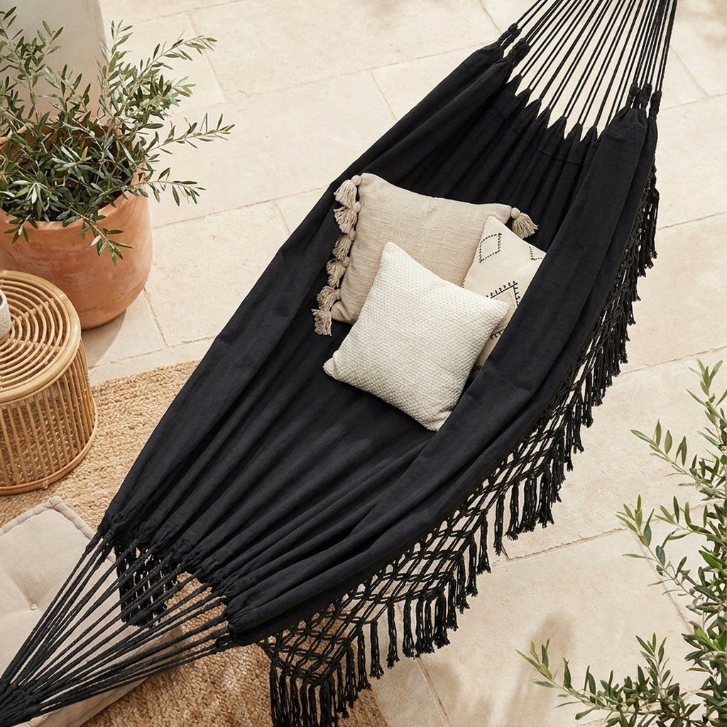 High-angle view looking down into a black fringe macrame hammock holding three cushions, above a woven jute rug on a stone tile patio with a rattan side table