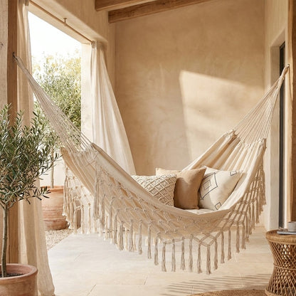 Cream fringe macrame hammock with a diamond netting border and long knotted fringe, holding three cushions, in a covered patio with wood ceiling beams, sheer curtains and a potted olive tree