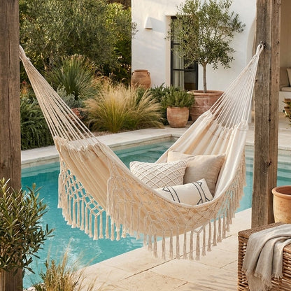 Cream fringe macrame hammock with three cushions hung between wood posts beside a turquoise swimming pool, with a white stucco building, olive trees and a wicker chair behind