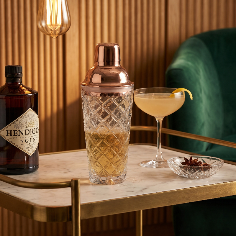 Glass cocktail shaker with copper lid on table beside wine bottle