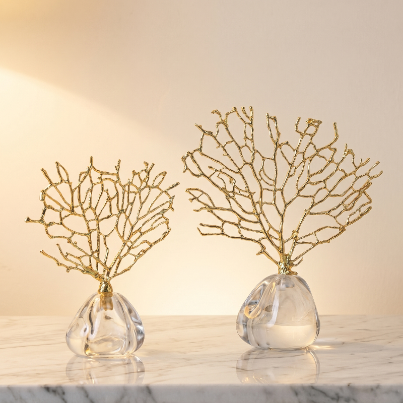 Two decorative gold branch sculptures in clear glass vases on a marble surface with a warm light background.