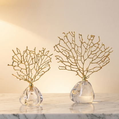 Two decorative gold branch sculptures in clear glass vases on a marble surface with a warm light background.