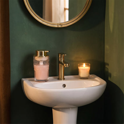 Clear glass soap dispenser with blush-pink soap and a gold pump on a white pedestal sink with a brass faucet and candle against a dark green wall